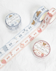 Two rolls of decorative washi tape with matching round spools on a white background. One roll is light blue with white lace and gold motifs, and the other is pale pink with white lace, pink bows, and gold motifs.