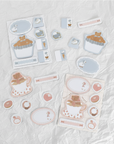 A set of cute, pastel-themed sticker sheets and individual stickers featuring a cupcake, coffee cup, and bunny motifs, arranged on a brown textured leather background.