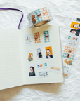 A roll of decorative washi tape and matching printed strips displayed on an open grid notebook. The tape features illustrated portraits, small photos, plaid patterns, and text labels in soft pastel tones.