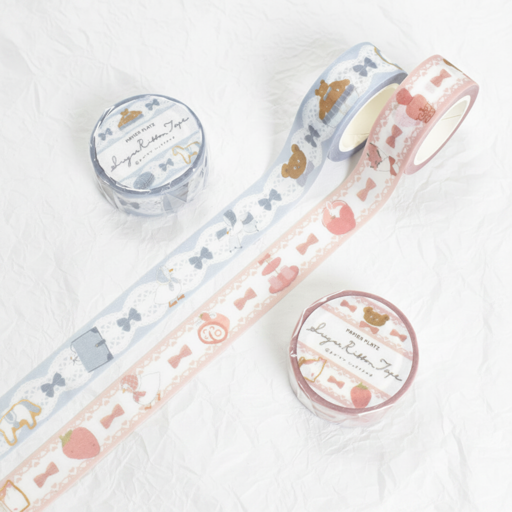 Two rolls of decorative washi tape with matching round spools on a white background. One roll is light blue with white lace and gold motifs, and the other is pale pink with white lace, pink bows, and gold motifs.
